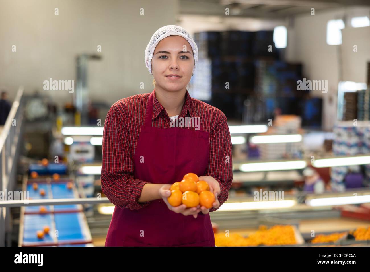 Giovane lavoratrice allegra e positiva in uniforme colorata tiene in mano tangerini freschi maturi vicino alla linea di trasporto. Foto Stock