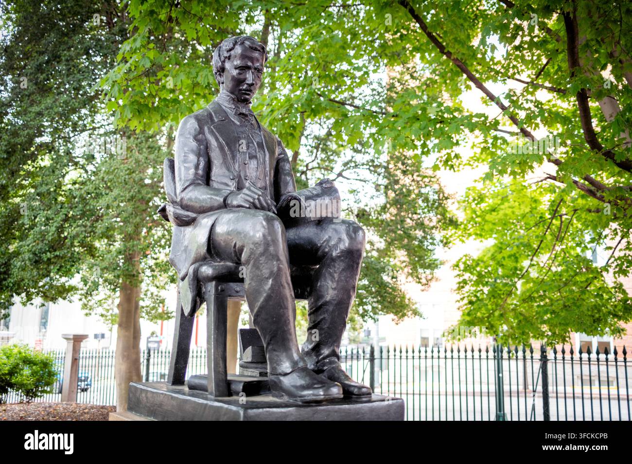 Statua di Poe dietro una recinzione in ferro a Richmond, Virginia | RICHMOND, Virginia — Recinzioni in ferro battuto e la cornice perimetrale della piazza incorniciano questa vista a tre quarti di Edgar Allan Poe, con una tavoletta appoggiata sul ginocchio. Il trattamento modernista dello scultore Charles Rudy rappresentava una rottura intenzionale con lo stile commemorativo precedente della piazza — la stessa convenzione del ritratto seduto usata per la statua di Hunter Holmes McGuire del 1904 nelle vicinanze, qui privata di qualsiasi gesto o emozione di stile Beaux-Arts. Foto Stock