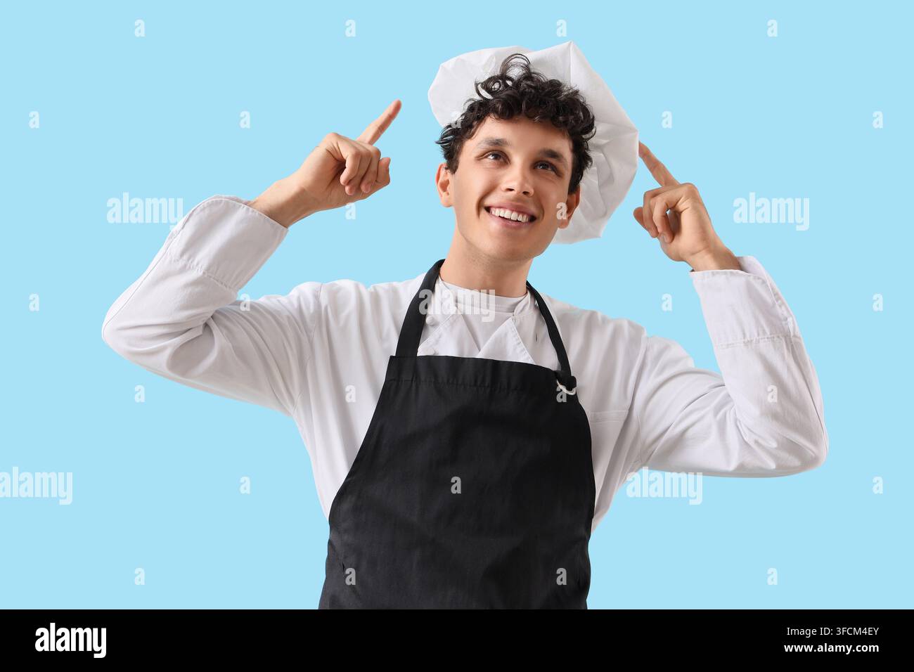 Ispirazione culinaria e cucina creativa, giovane chef che indica verso lo spazio di copia con un sorriso in uniforme professionale su uno sfondo blu chiaro Foto Stock