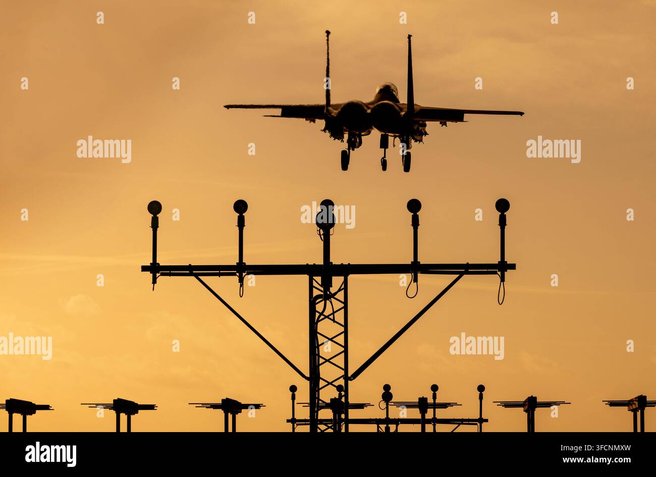 Jet da combattimento F-15 che si avvicina per atterrare al tramonto, silouettato contro un cielo arancione con le luci di avvicinamento alla pista in primo piano. Foto Stock