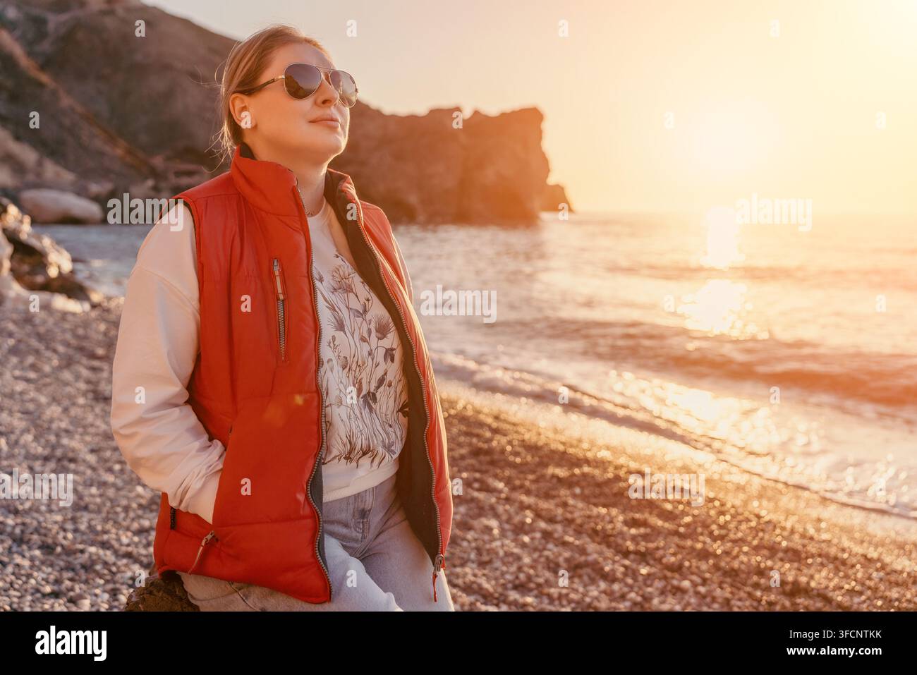 Donna tramonto spiaggia donna di mezza età indossa un giubbotto rosso e occhiali da sole si trova su una riva rocciosa godendosi lo scenario dell'ora dorata dell'oceano Foto Stock