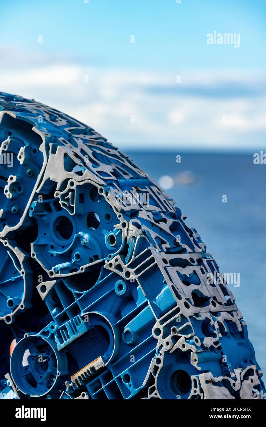 una grande scultura circolare realizzata con parti meccaniche blu e grigie stratificate, ambientata su uno sfondo sfocato di mare e cielo Foto Stock