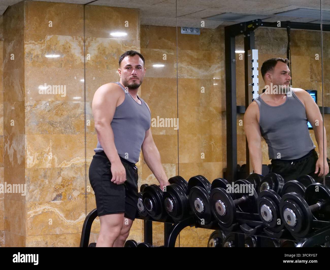 Uomo muscoloso in piedi vicino a una rastrelliera di manubri in una palestra di alto livello, che guarda in una grande parete a specchio. Si sta preparando per un allenamento di forza se Foto Stock