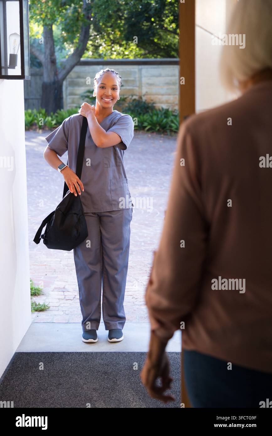 Infermiera e donna anziana in piedi alla porta di casa con camici grigi che tengono una borsa Foto Stock