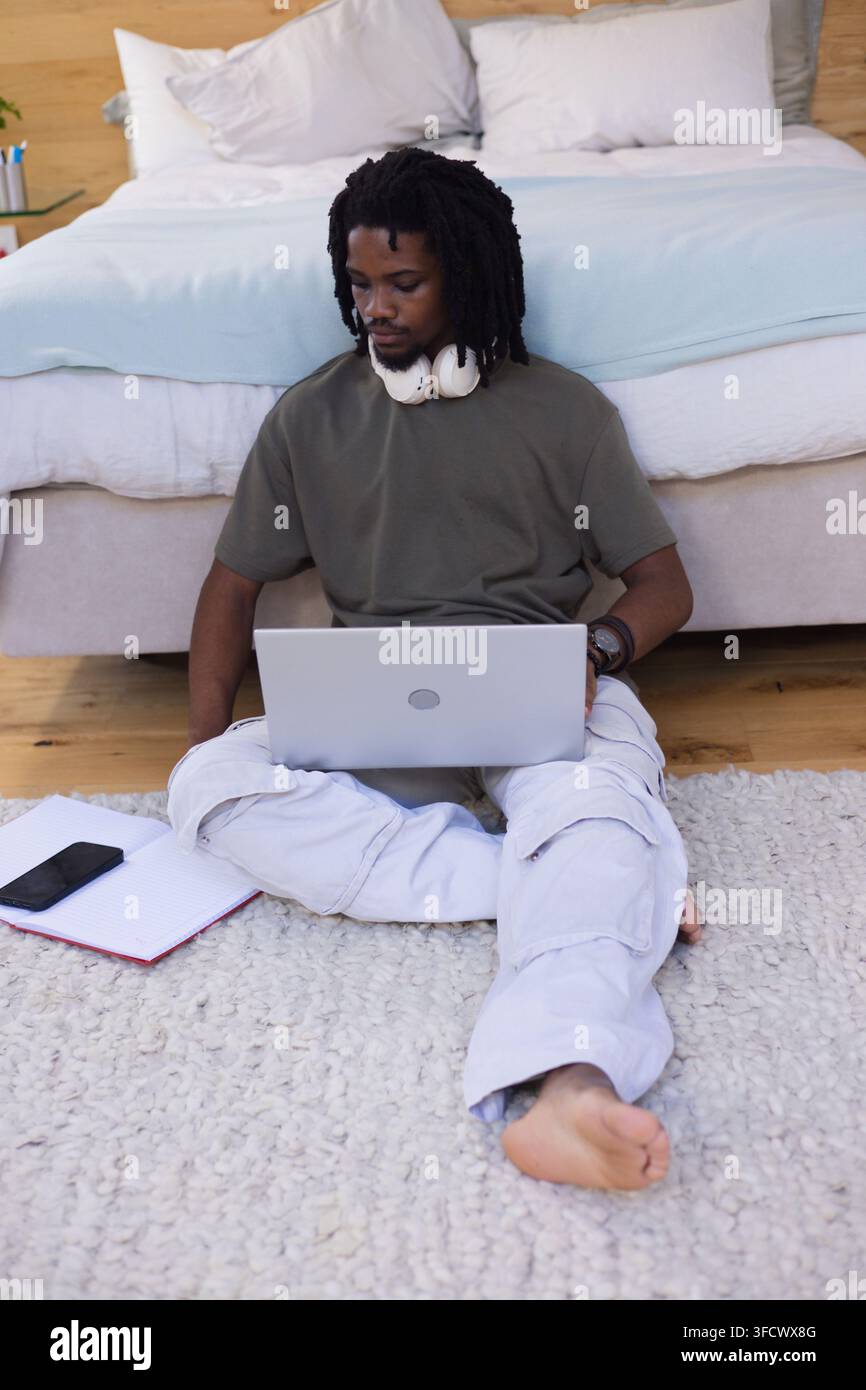Uomo sulla ventina seduto su un tappeto in camera da letto che usa un laptop con cuffie, smartphone, quaderno nelle vicinanze Foto Stock