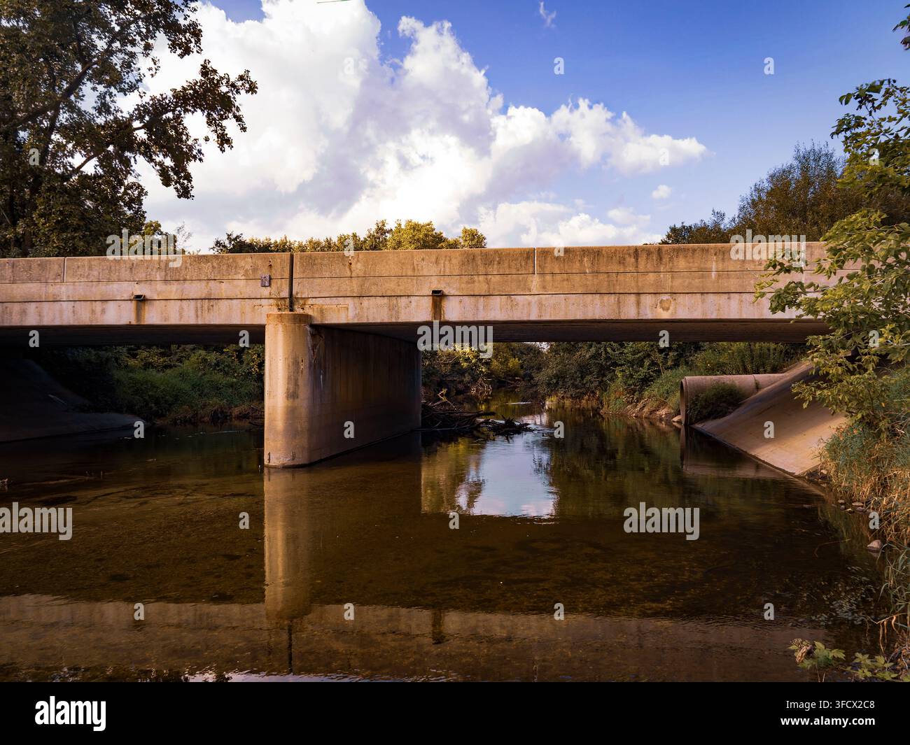 Ponte di cemento su un fiume calmo che scorre attraverso un paesaggio verdeggiante con soffici nuvole bianche nel cielo azzurro Foto Stock