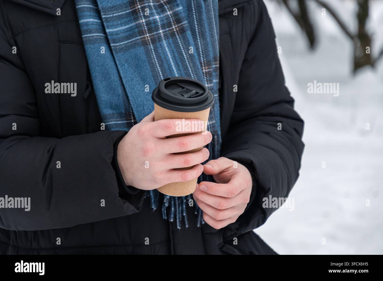 Primo piano di un uomo che tiene una tazza di caffè da asporto con entrambe le mani all'aperto in un parco innevato in inverno. Concetto di bevanda calda, tempo freddo e stile di vita invernale. Foto Stock