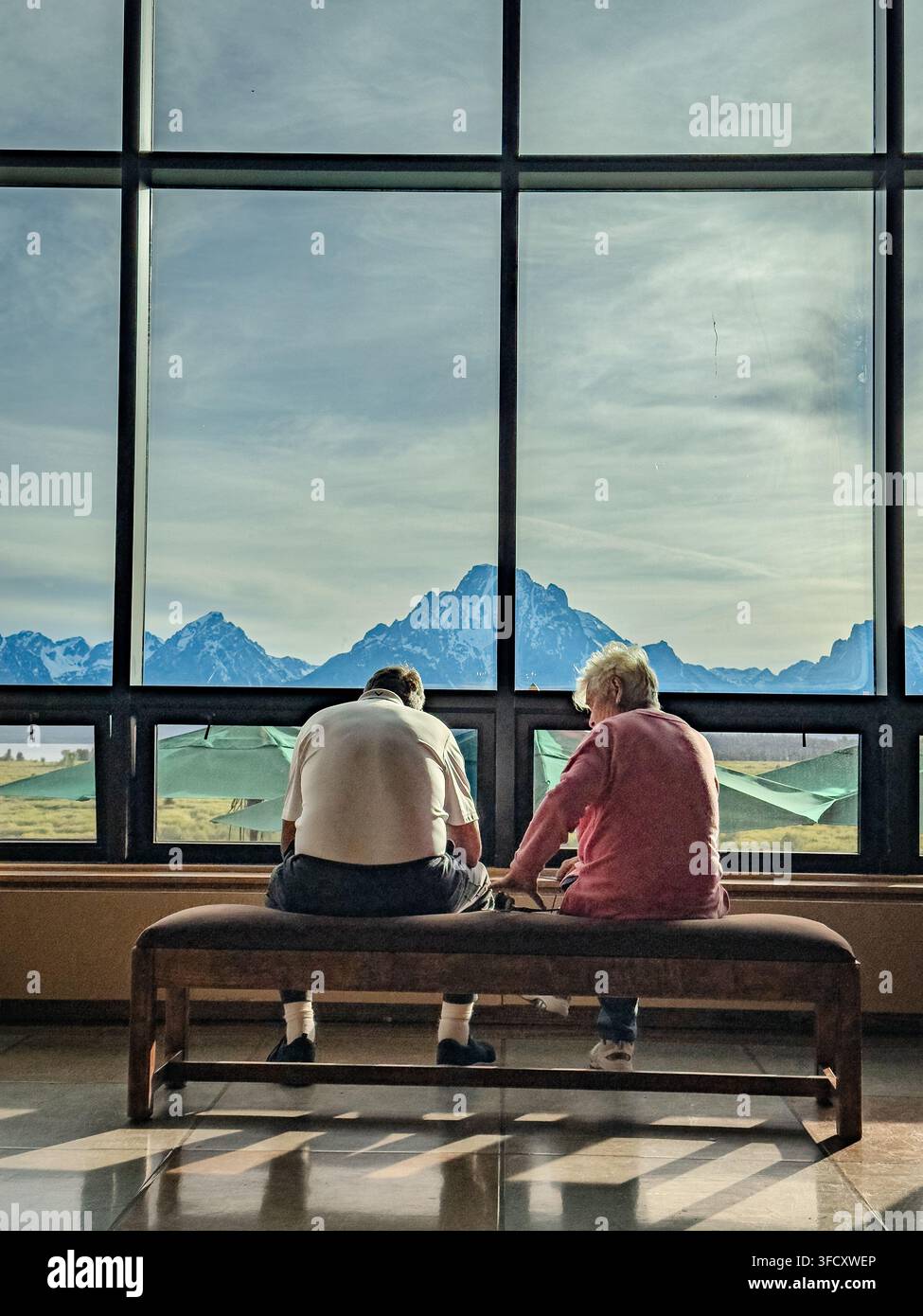 Due visitatori sono seduti su una panchina all'interno del Jackson Lake Lodge, incorniciati da finestre a tutta altezza e dalle vette lontane della catena del Grand Teton. Foto Stock