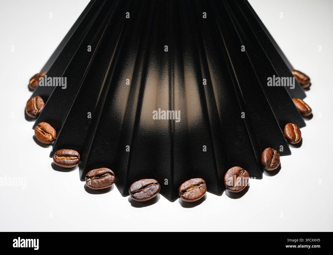 Close-up di chicchi di caffè tostati disposti intorno a un dripper da caffè in origami nero su una superficie bianca, scatto in studio. Foto Stock