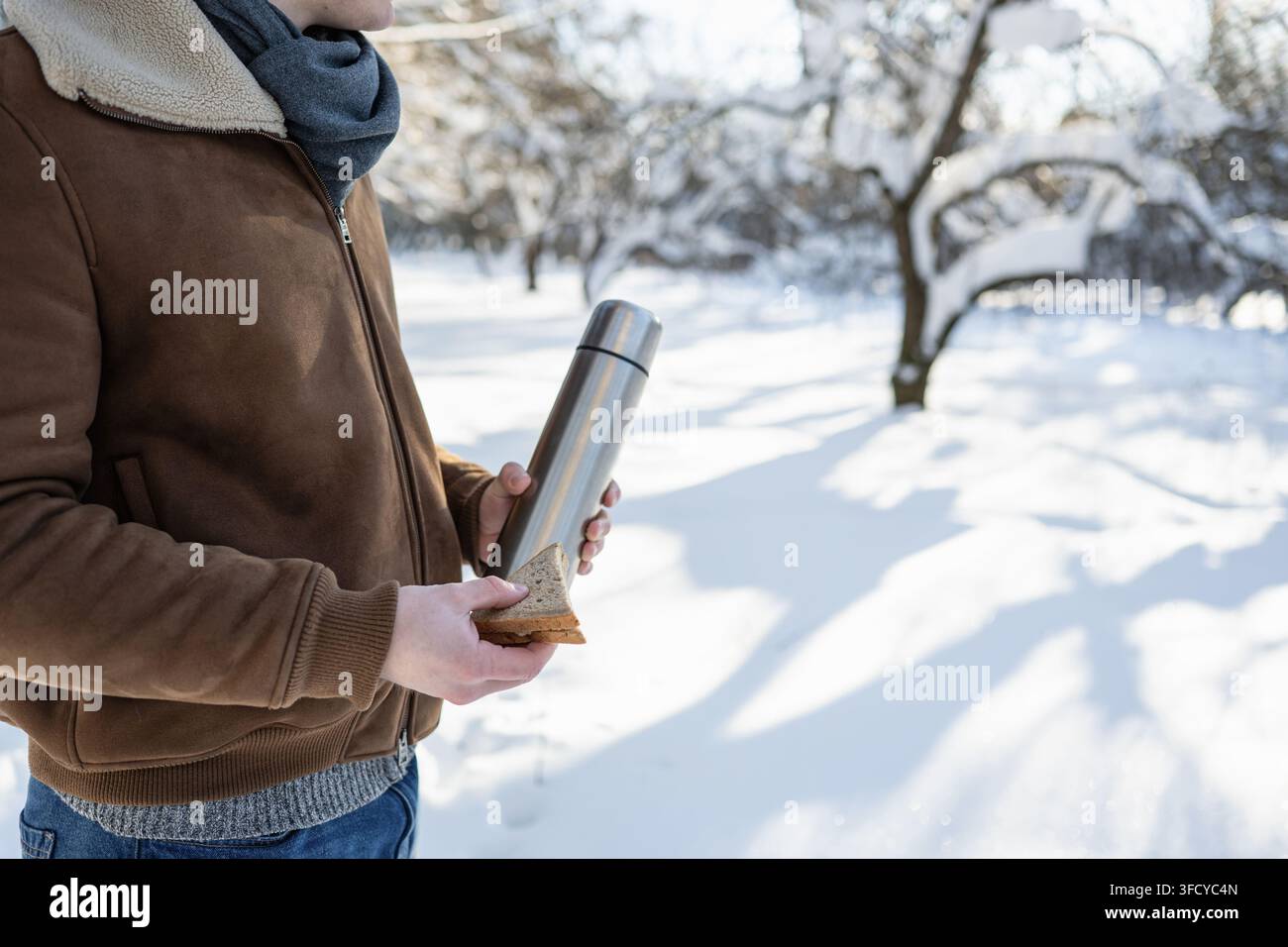 Giovane uomo che tiene una thermos di metallo e un panino in un parco innevato. Pranzo all'aperto invernale, bevanda calda e pausa con il freddo, concetto con spazio per il testo. Foto Stock