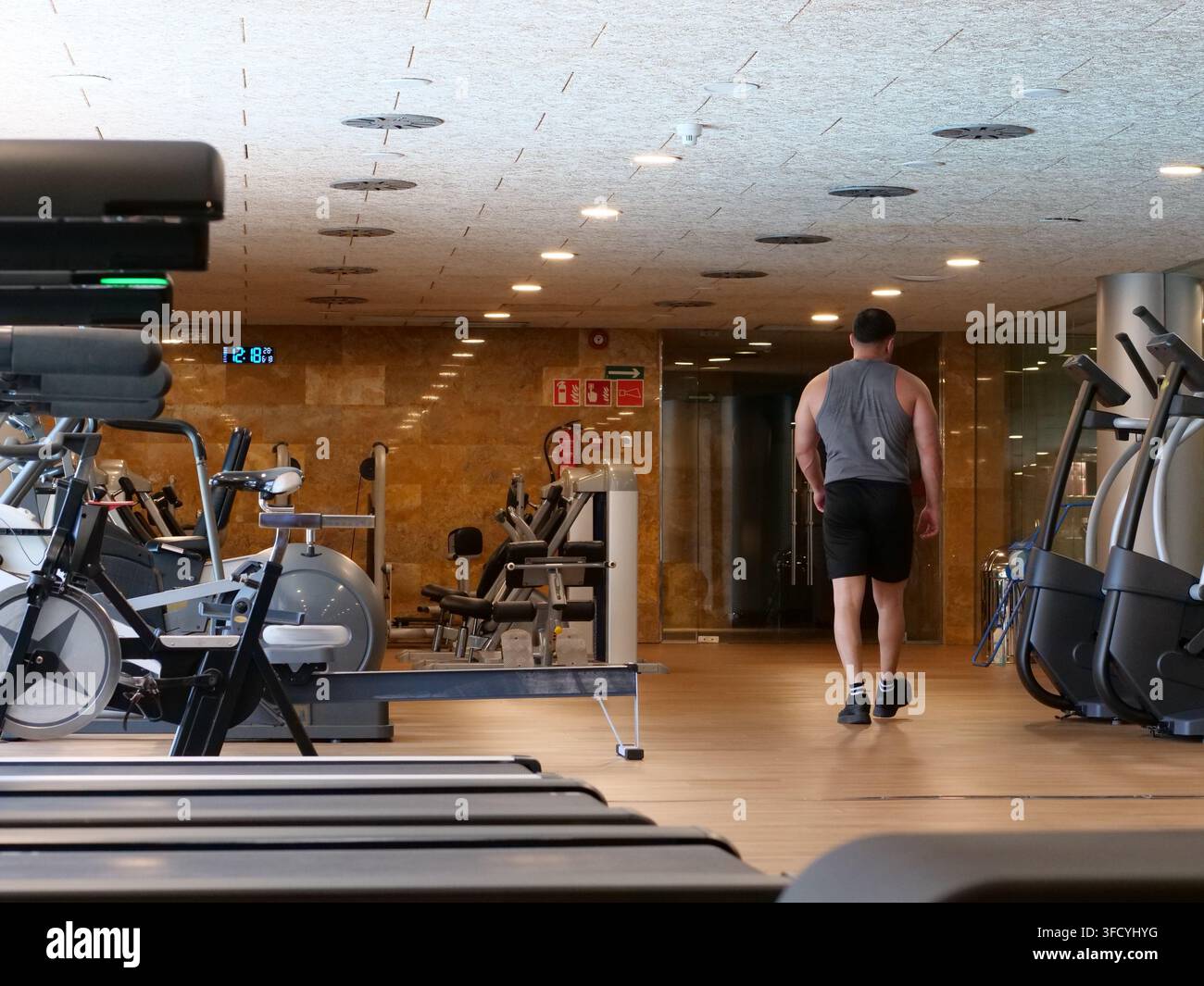 Vista posteriore di una figura maschile atletica che si allontana attraverso un corridoio vuoto di una moderna palestra. Circondato da apparecchiature cardio professionali, tapis roulant e resi Foto Stock