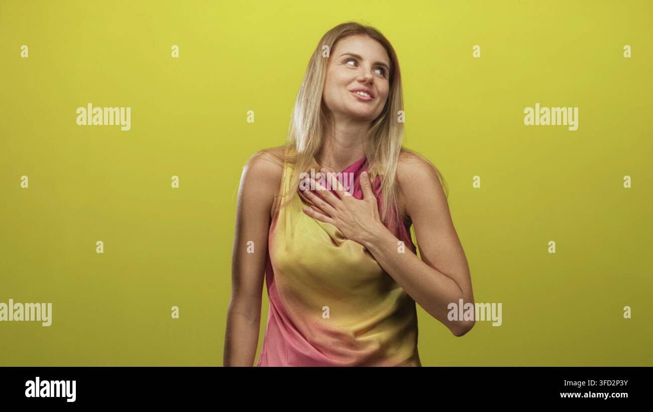 La donna bionda mette la mano sul petto in uno sfondo giallo da studio, sorridendo con uno sguardo attento verso l'alto; gratitudine calore sincerità. Foto Stock