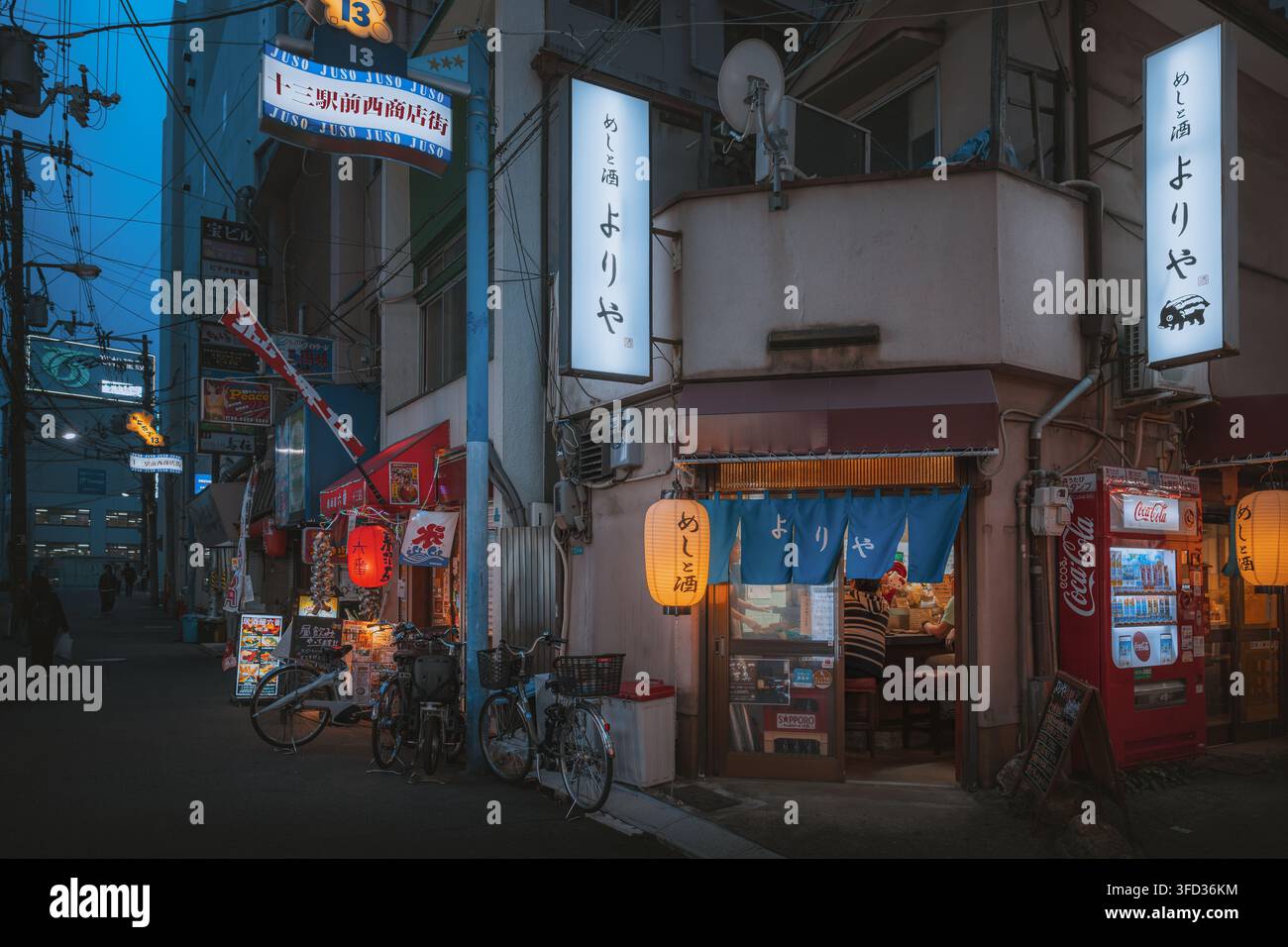 Osaka, Giappone - 26 aprile 2026: Vista dell'ingresso di un ristorante giapponese tradizionale con lanterne luminose, tende noren blu e biciclette parcheggiate su un Foto Stock