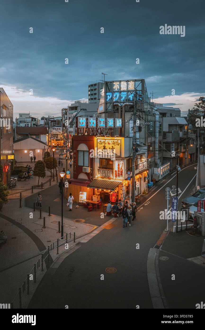 Giappone - 03 maggio 2026: Vista di una strada stretta al crepuscolo con insegne al neon luminose e persone che cenano all'esterno di un ristorante izakaya tradizionale. Foto Stock
