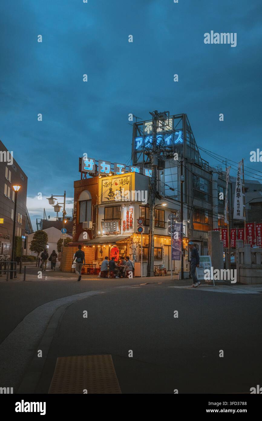Giappone – 03 maggio 2026: Vista di un piccolo ristorante all’angolo di una strada con persone che cenano all’aperto sotto insegne al neon luminose al crepuscolo. Foto Stock