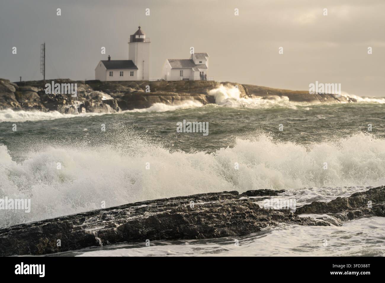 Il contrasto soleggiato della tempesta porta luce calda al faro di Langoytangen mentre un'onda grande in primo piano si infrange su scogli scuri in Norvegia per un tempo drammatico Foto Stock