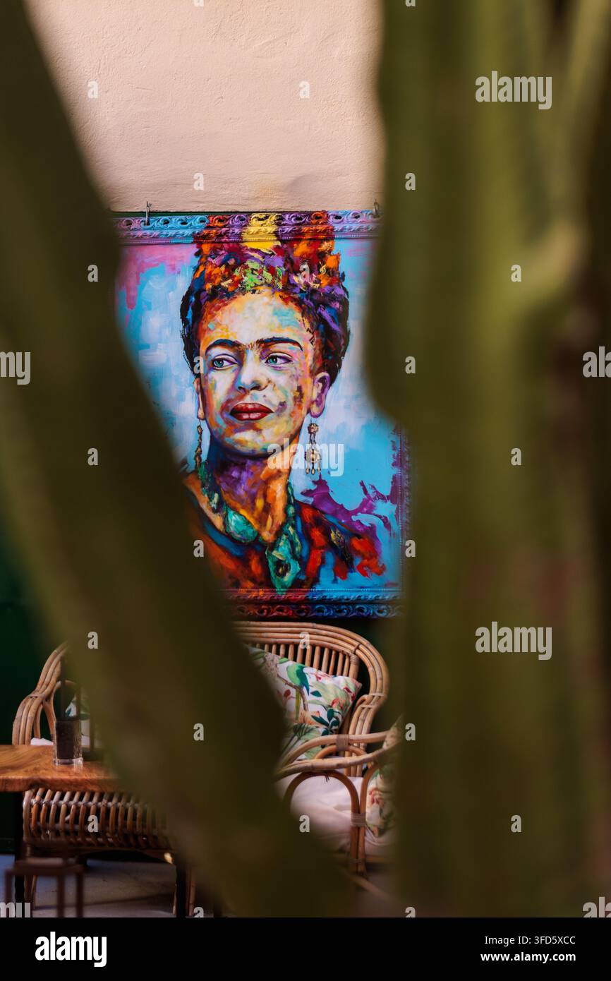 Un dipinto ritratto colorato dell'iconica artista messicana Frida Kahlo appeso a un muro di un patio all'aperto, scattato attraverso una cornice naturale. Foto Stock