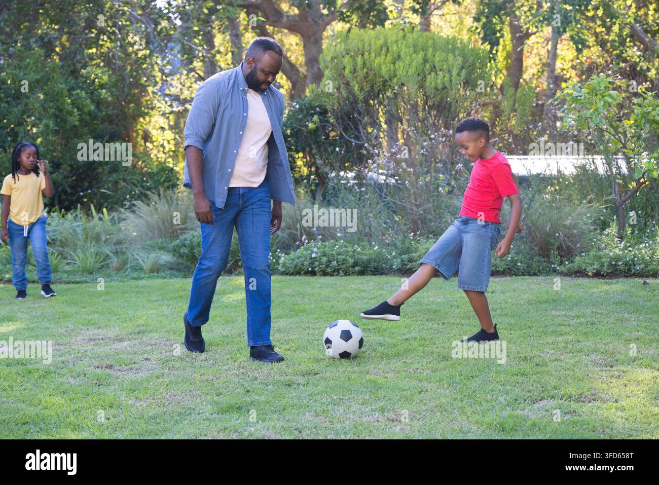 Padre con due bambini che giocano con un pallone da calcio sul prato del giardino, il padre indossa una camicia azzurra Foto Stock