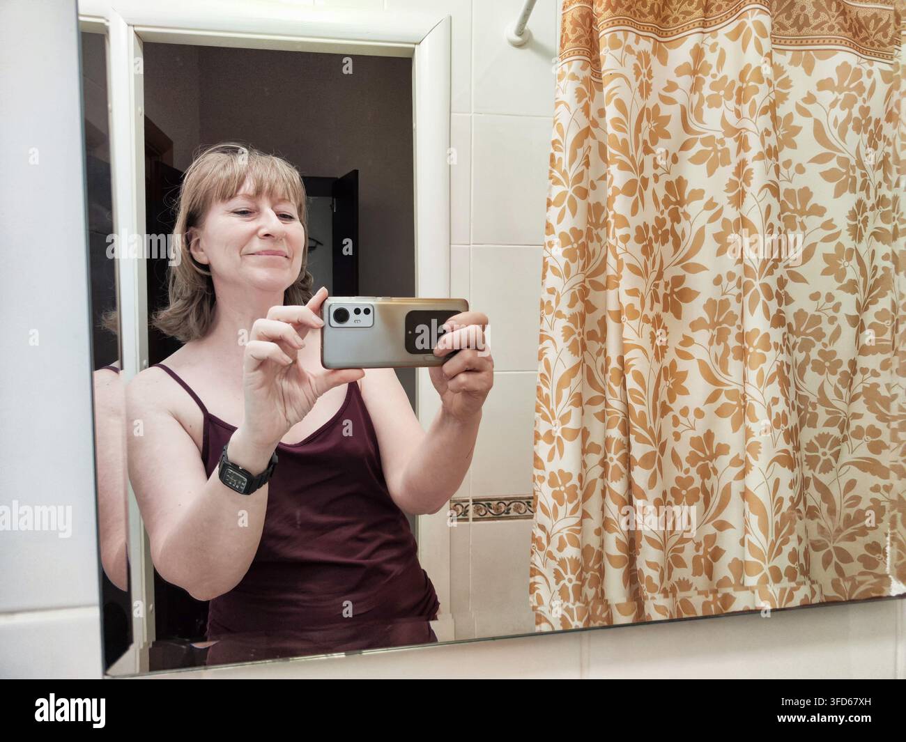 Una donna si trova davanti a uno specchio nel suo bagno e scatta un selfie con uno smartphone. Sorride e si sente a suo agio nell'ambiente che la circonda. Foto Stock