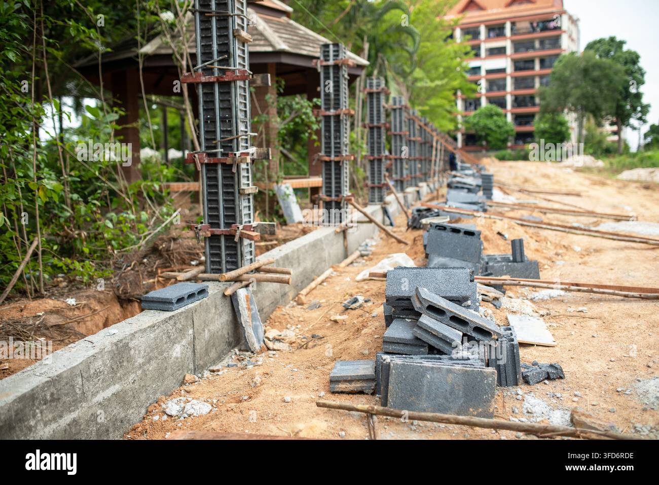 Pali di recinzione in cemento rinforzato si allineano lungo una fondazione preparata, blocchi di cemento cavi impilati attendono l'installazione, un edificio residenziale si erge oltre Foto Stock