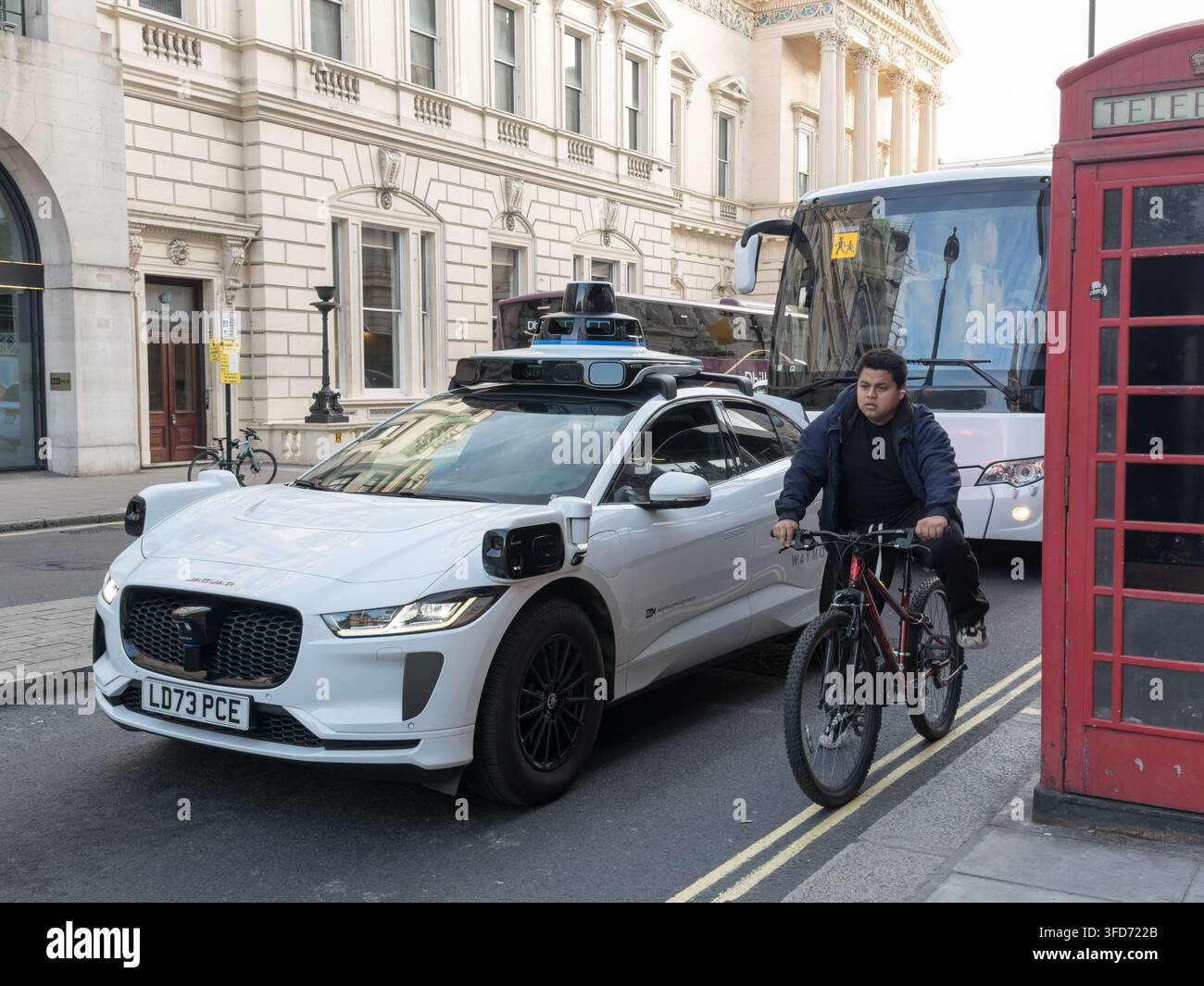 Un ciclista passa tra un Robotaxi autonomo Waymo, visto dal lato del passeggero anteriore, e una cabina telefonica rossa visibile a Londra Foto Stock