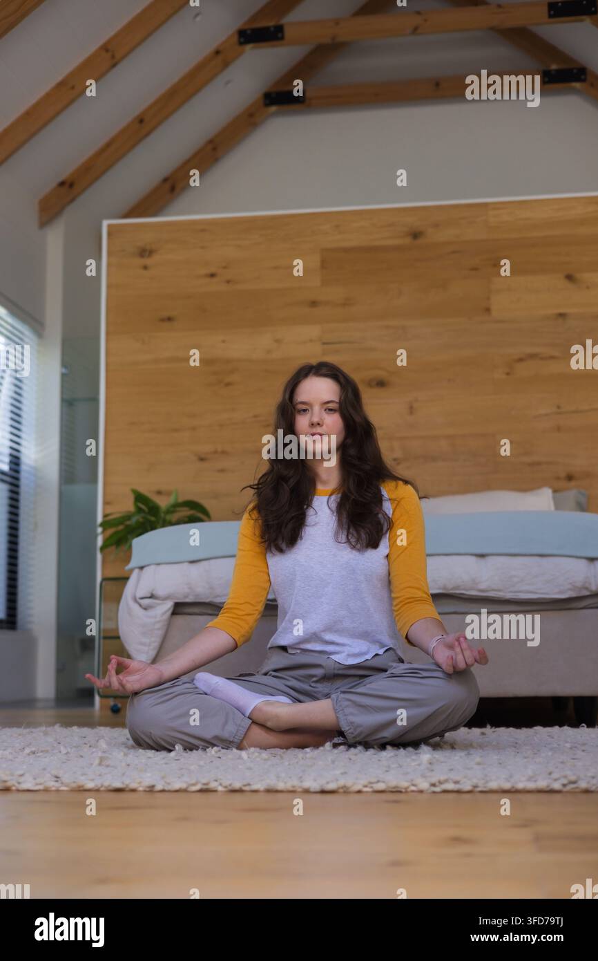 Donna seduta a gambe incrociate in meditazione su un tappeto in camera da letto, indossa una maglietta raglan con maniche gialle Foto Stock