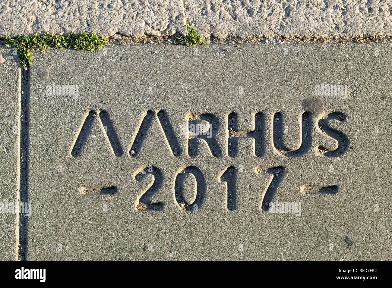 Primo piano di una lastra di marciapiede in cemento stampata con la parola Aarhus, che è la seconda città più grande della Danimarca, con piccolo muschio verde che cresce lungo th Foto Stock