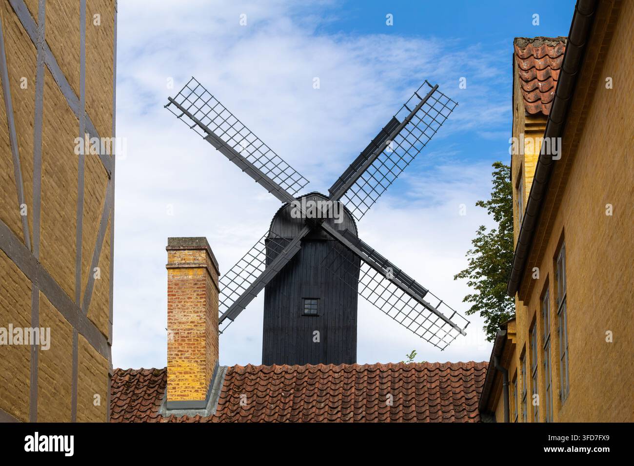 Tradizionale mulino a vento in legno nero che si erge sopra case in mattoni gialli con tetti di tegole rosse sotto un cielo blu luminoso. Foto Stock