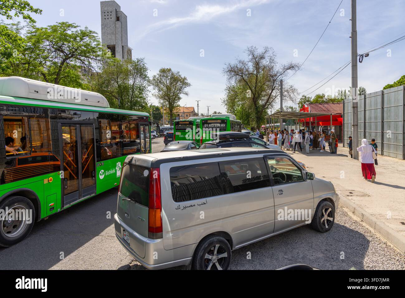 Biscikek, Kirghizistan - 6 maggio 2025: Una scena di strada con autobus pubblici verdi e un minivan argentato a una fermata dell'autobus. Le persone si radunano sotto un rifugio con tetto rosso Foto Stock