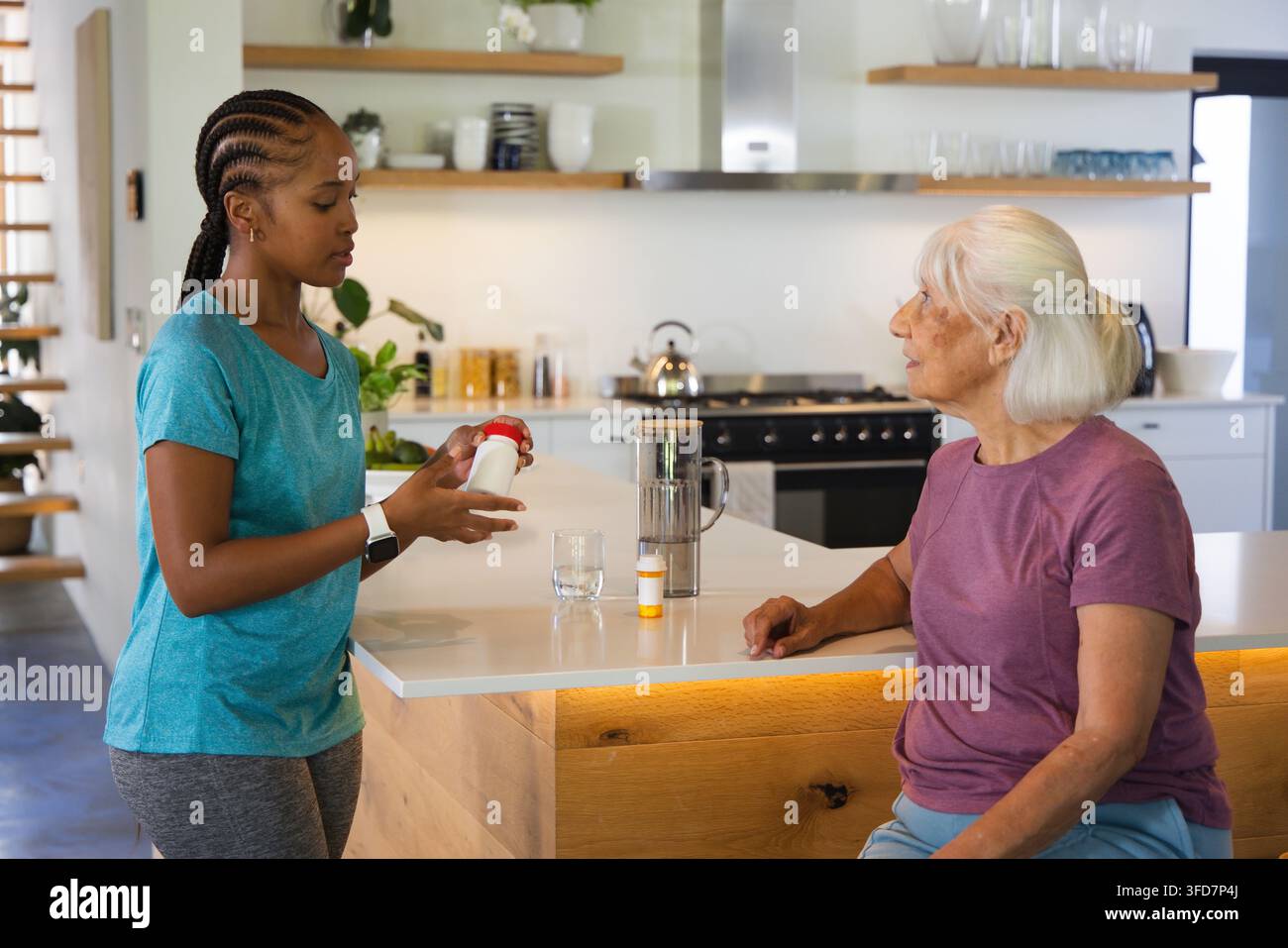 Caregiver afroamericana con orologio che consegna una bottiglia con tappo rosso a una donna anziana al bancone della cucina Foto Stock