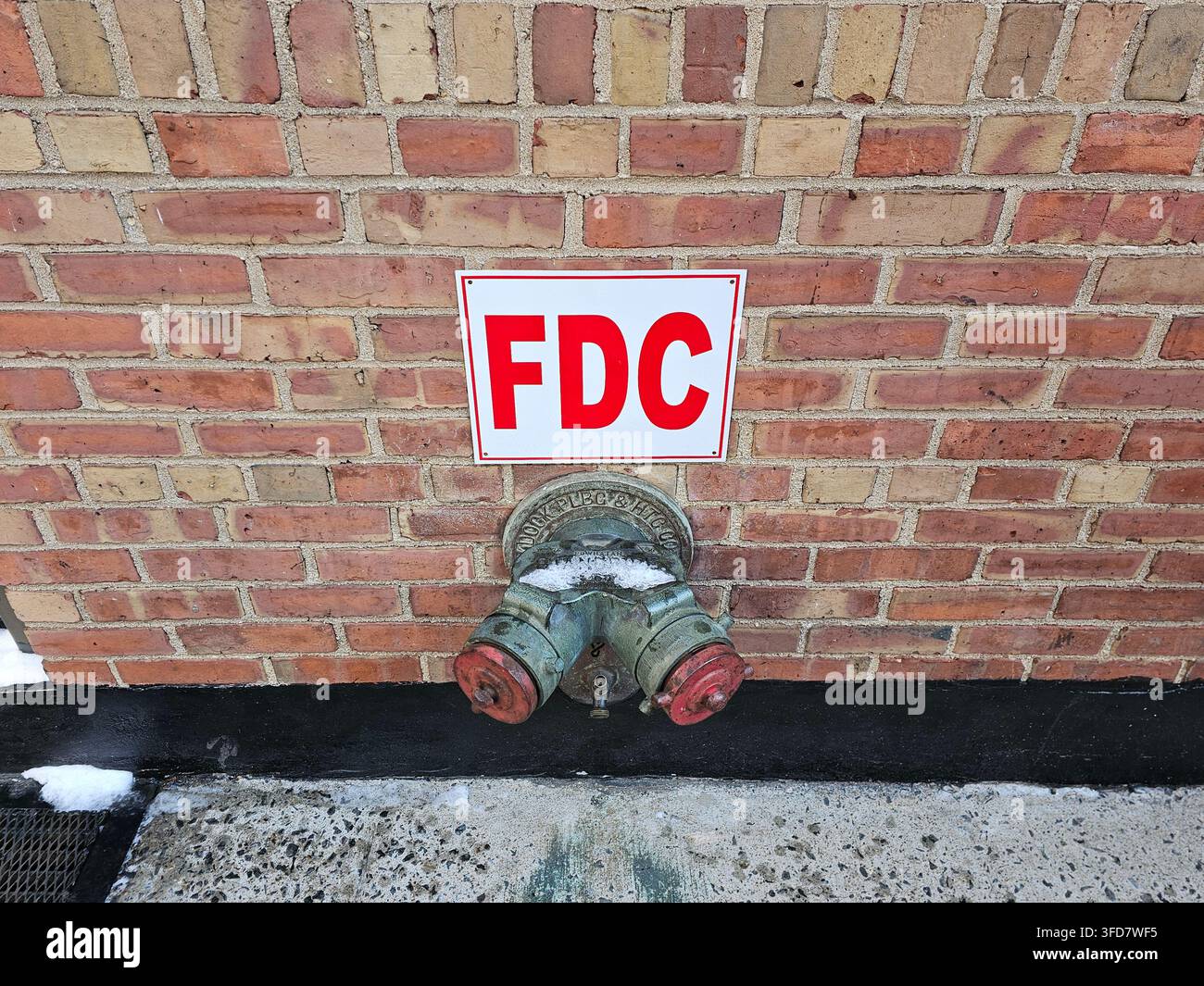 Segnale FDC rosso su un muro di mattoni sopra un raccordo antincendio siamese con due uscite tappate e catene, pannello nero alla base del muro in inverno. - Immagine stock catturata con smartphone