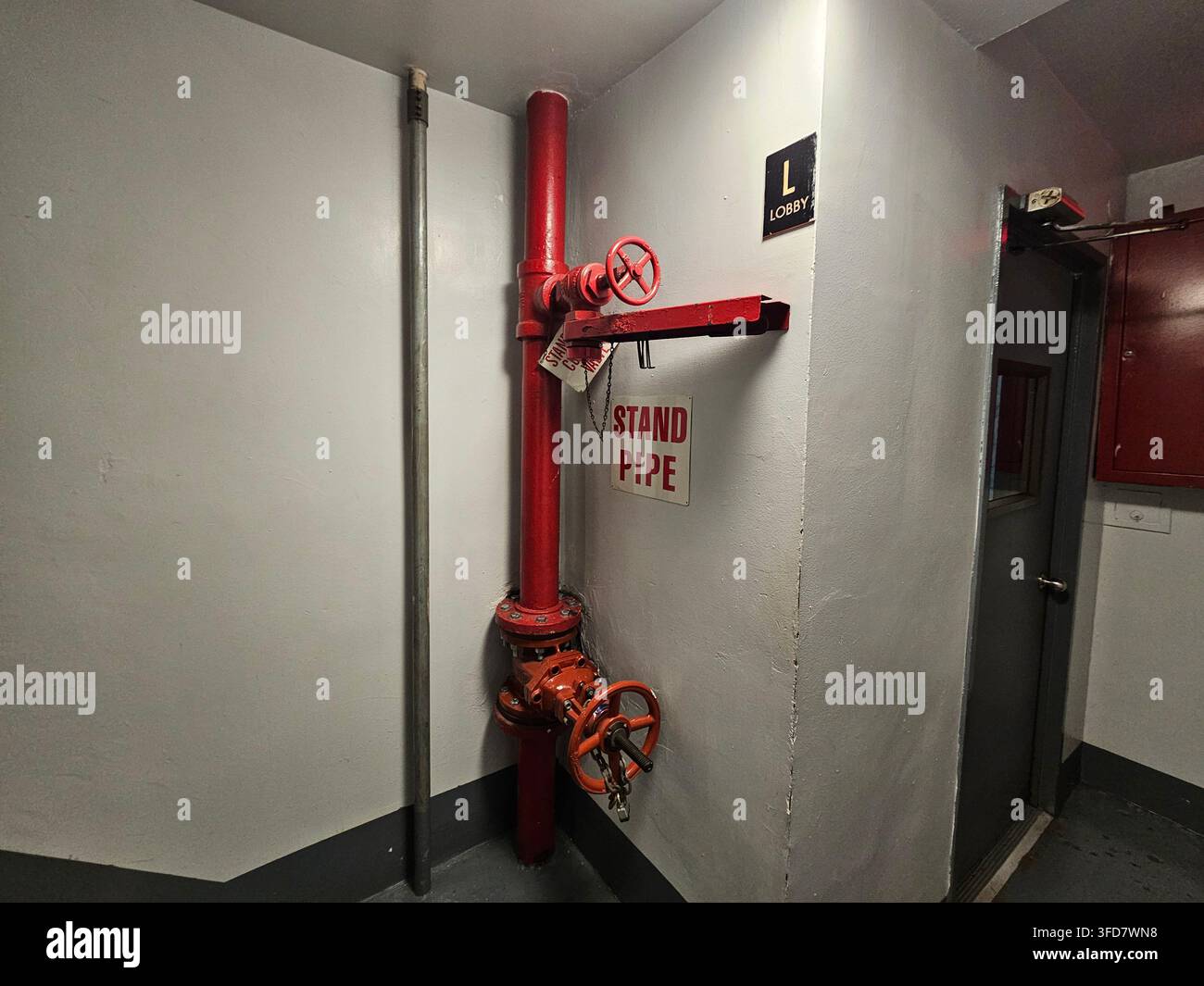 Tubo di risalita rosso con ruote di valvola all'interno di una tromba delle scale di un edificio commerciale, cartello STAND PIPE montato a parete accanto e porta scura sullo sfondo. - Immagine stock catturata con smartphone
