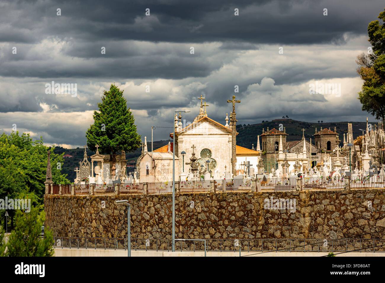 Lamego Portogallo, Vecchia chiesa con muro di pietra, cielo nuvoloso sopra. Gli alberi si ergono alti accanto a un antico cimitero. nella scena. L’atmosfera è solenne, suggestiva, storica Foto Stock