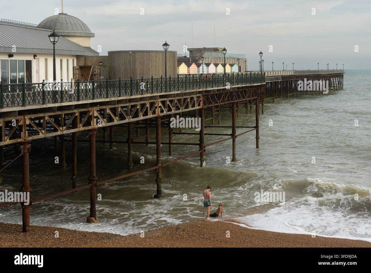 Due giovani uomini si riposano sulla riva ghiaiosa sotto il storico Hastings Pier nell'East Sussex, godendosi una giornata costiera al mare. Foto Stock