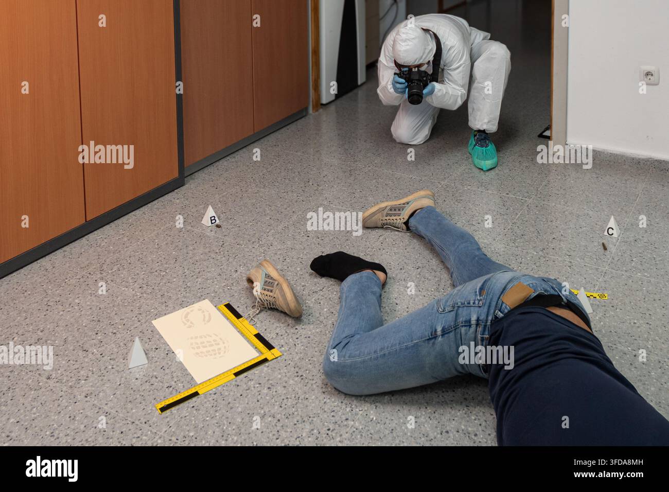 Investigatore forense inginocchiato per fotografare prove contrassegnate vicino a una vittima simulata, con una scala di misurazione visibile sul pavimento interno. Foto Stock