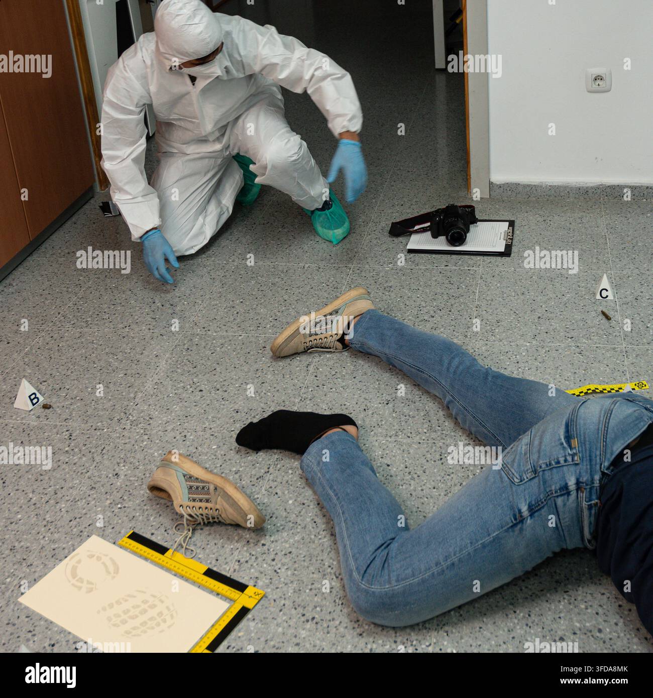 Investigatore forense esamina prove marcate sul pavimento vicino a una vittima simulata, con fotocamera, blocco note e scala di misurazione disposti per la documentazione. Foto Stock