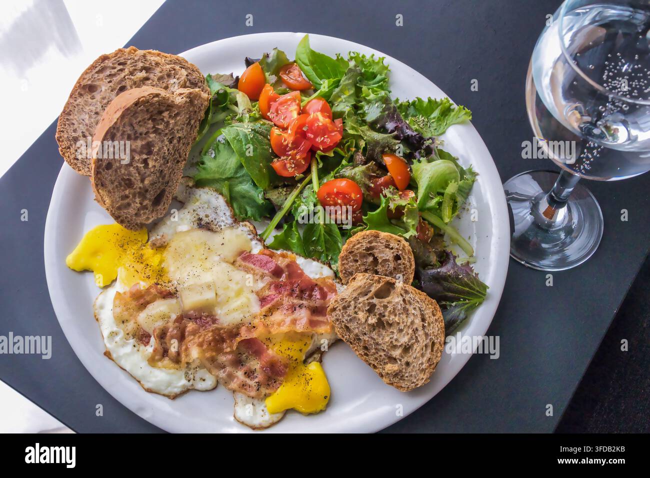 Una colazione deliziosa e bilanciata servita su un piatto bianco su un tavolo scuro. Il pasto presenta uova fritte ricoperte di formaggio fuso e pancetta croccante, un Foto Stock