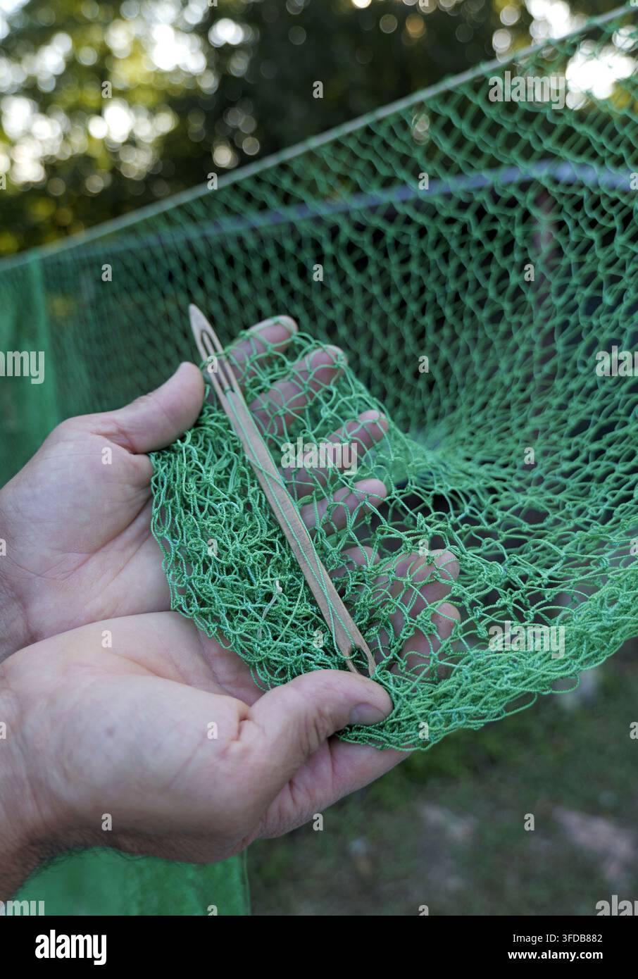 Mani che tessono una rete da pesca verde con una navetta di legno, primo piano che mostra una tecnica di annodatura tradizionale, artigianato abile e paziente uomo manuale all'aperto Foto Stock