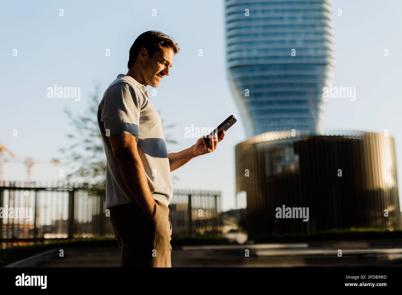 Un uomo si trova rilassato nel paesaggio urbano, controllando il suo smartphone mentre il sole tramonta dietro un edificio moderno. Foto Stock