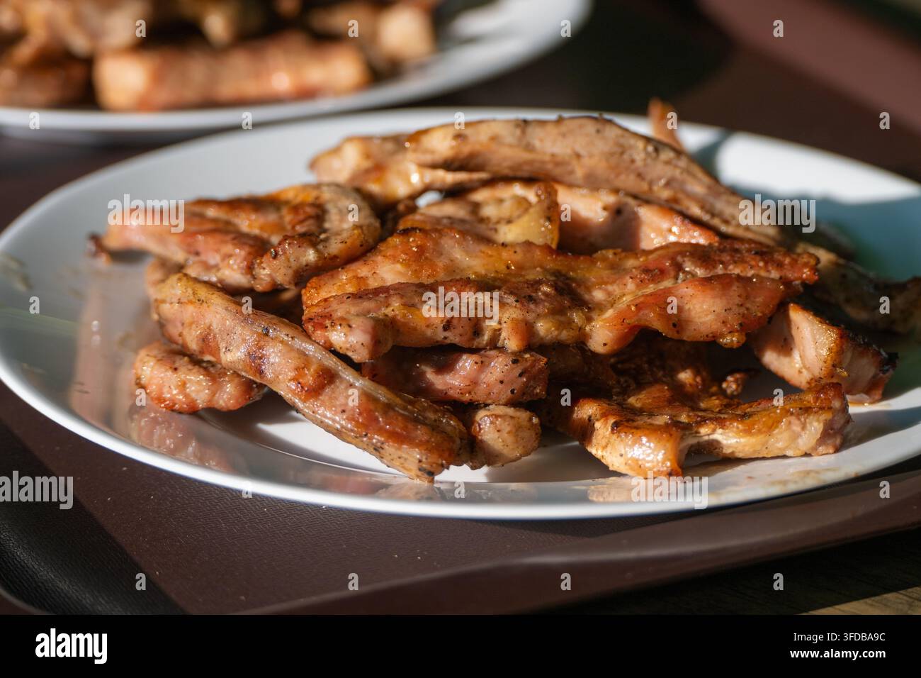 Un piatto bianco colmo di succose costine e strisce di pancetta di maiale appena grigliate, condite con pepe nero macinato, alla luce naturale del sole. Foto Stock