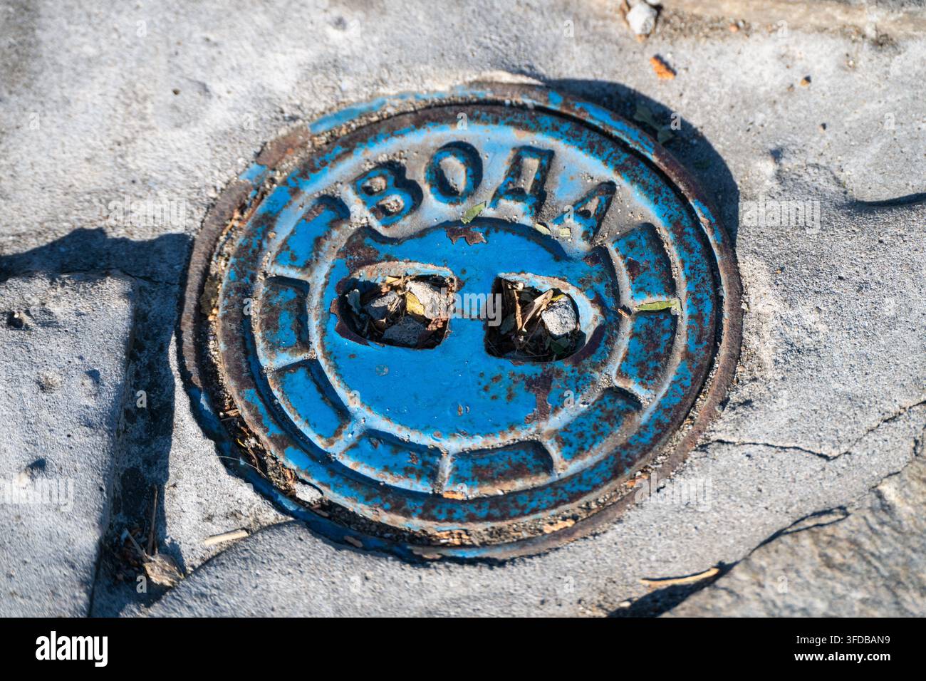 Un tappo circolare blu in ghisa con testo cirillico per l'acqua è incastonato in un marciapiede di cemento. Foto Stock