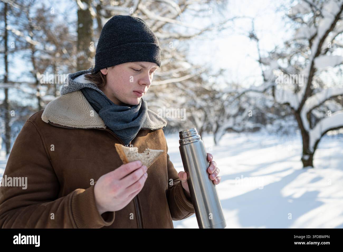 Giovane uomo che beve tè caldo e mangia un panino nel parco invernale Foto Stock