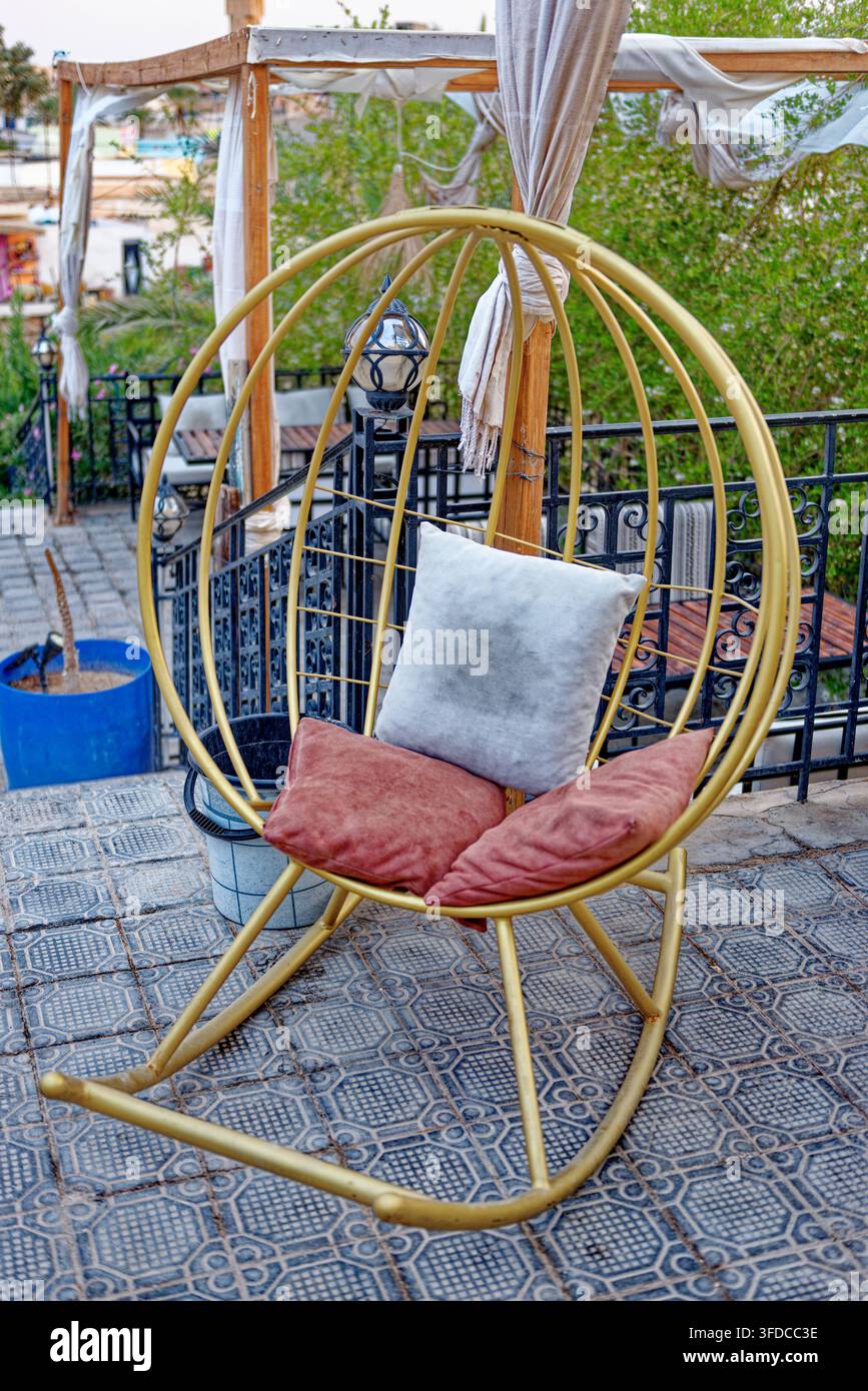 Sedia a dondolo sferica in filo metallico tubolare dorato realizzata a mano con cuscini rossi e grigi su una terrazza piastrellata all'aperto ad Hammat-al-Jarid. Cattura Foto Stock