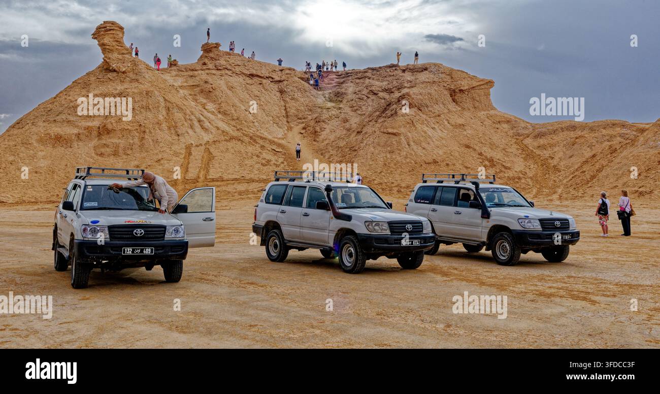 Tre SUV 4x4 bianchi parcheggiati nelle pianure desertiche sotto l'iconica formazione rocciosa Camel Rock con turisti che scalano la vetta a Ong Jmal, Tunisia. Catturato Foto Stock