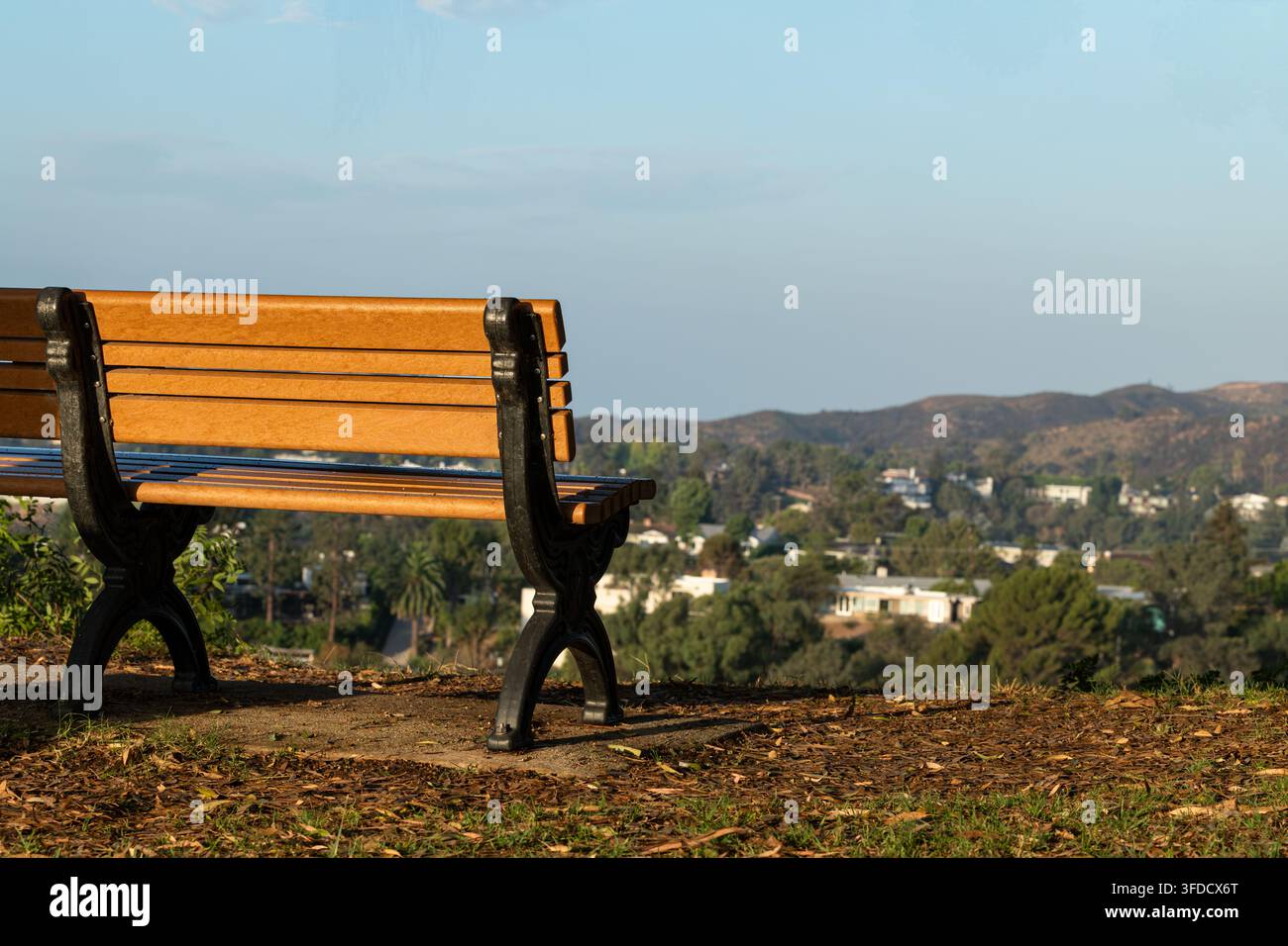 Panchina di legno nel parco che si affaccia su un paesaggio montano scenografico e una valle residenziale sotto un cielo blu soffice, creando un ambiente esterno tranquillo. Foto Stock