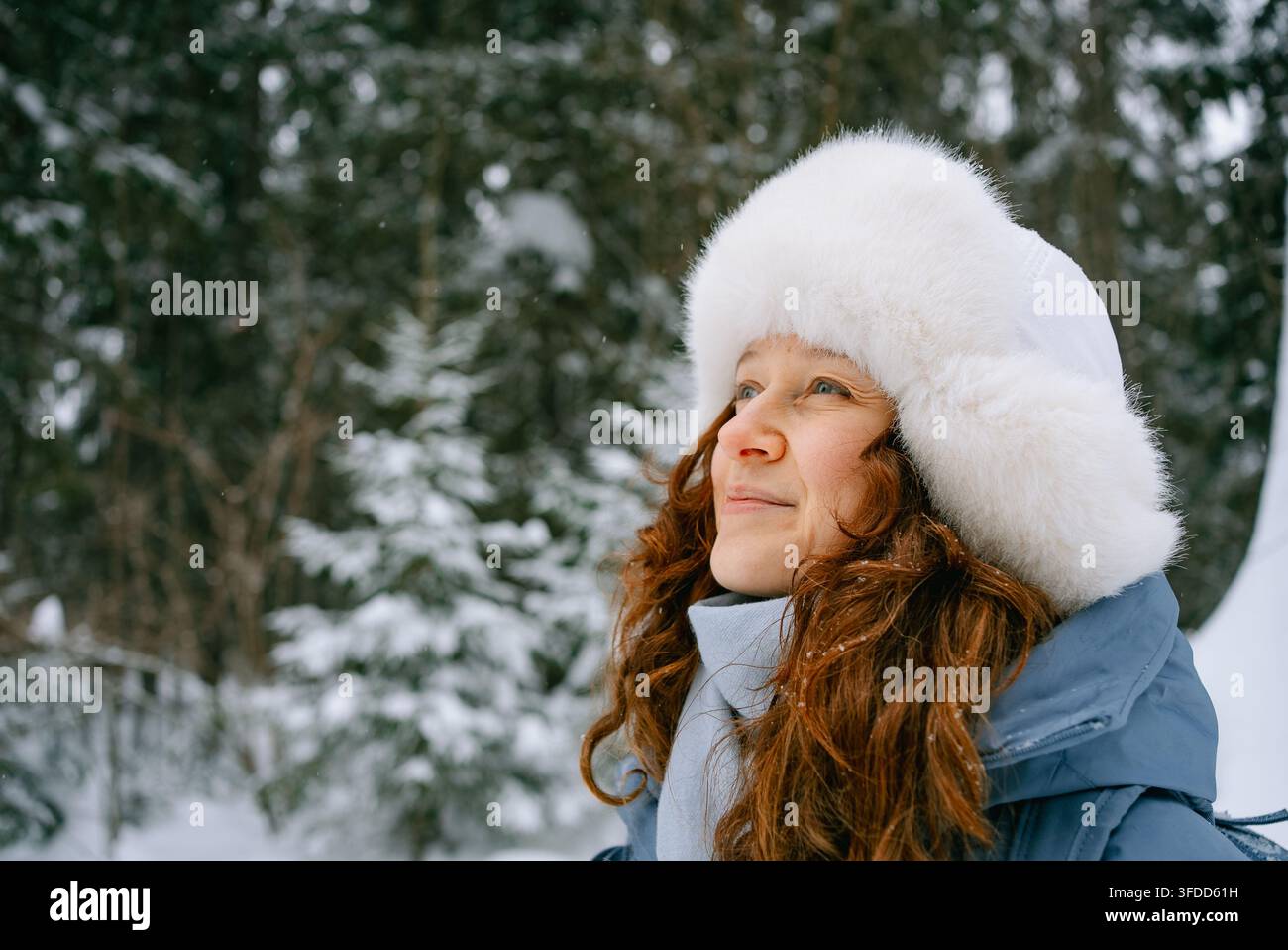 Donna con capelli rossi ondulati e cappello di pelliccia bianca che guarda verso l'alto e sorride in una foresta innevata d'inverno, godendosi il freddo e l'aria frizzante mentre esplora la natura Foto Stock