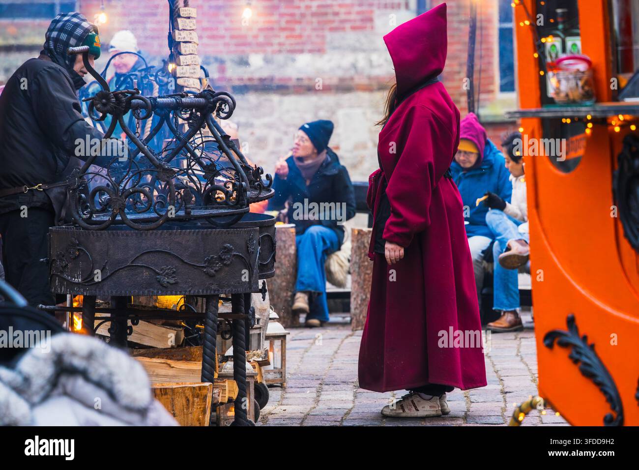 Una performance artigianale di mercato porta una persona con un cappuccio rosso accanto a attrezzature in ferro ornate e visitatori seduti al mercato natalizio di Riga per una tradizione vivente Foto Stock