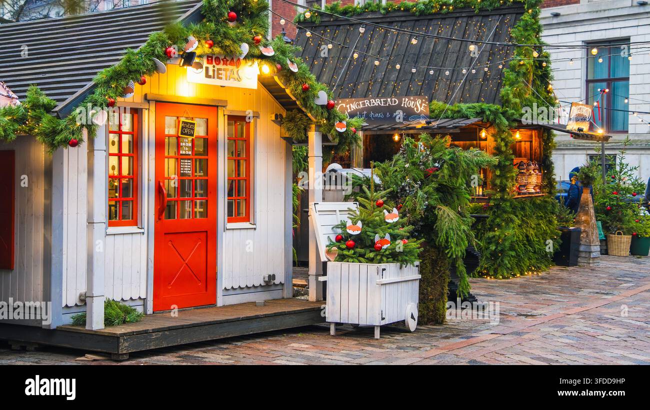 L'accoglienza decorata della cabina brilla attraverso porte rosse e ghirlande su una piccola capanna di legno al mercato di Natale di Riga per un'atmosfera fatta a mano e ospitalità invernale Foto Stock