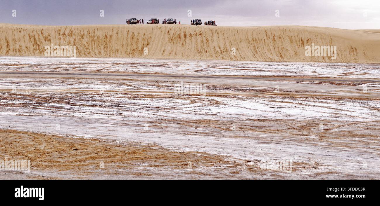 Filea di veicoli fuoristrada 4x4 e turisti parcheggiati sulla cresta di una massiccia duna di sabbia che si affaccia sulle pianure di sale bianche di Hammat-al-Jarid. Catturato Foto Stock