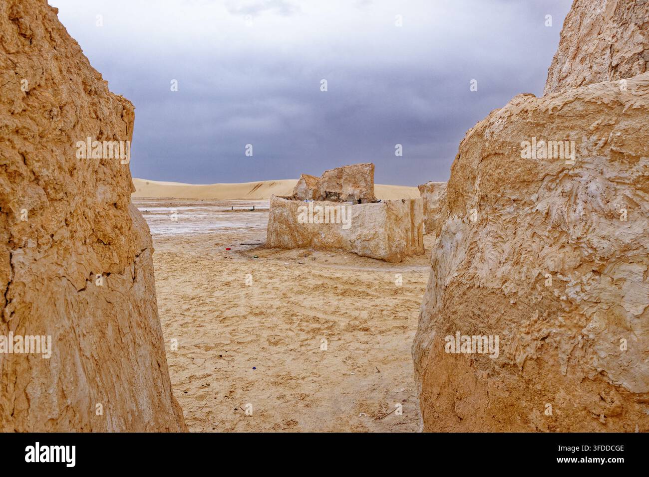 Vista attraverso muri di fango strutturati verso dune di sabbia e strutture in gesso autonome sotto un cielo tempestoso scuro a Mos Espa nell'Hammat-al-Jarid. Cap Foto Stock